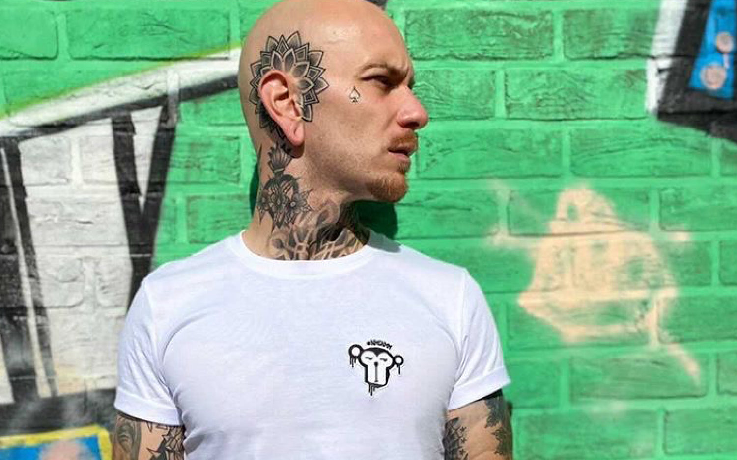 Mann vor grüner Wand mit weißem T-Shirt Aufnäher Affe auf der Brust